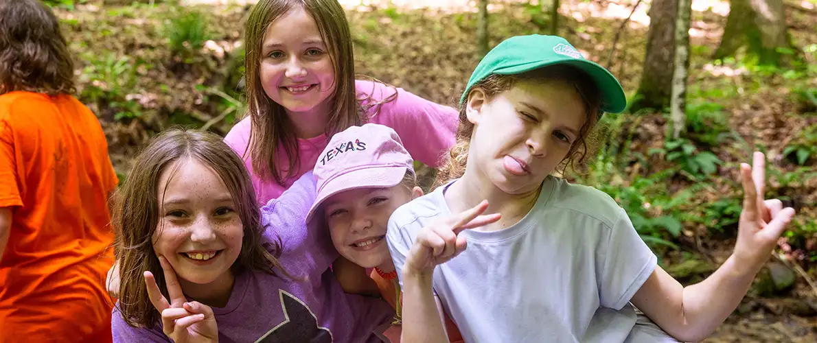 Four children smile and pose with peace signs while sitting outdoors in a wooded area during a summer camp activity.